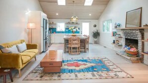 A spacious living room with a yellow sofa, a wooden coffee table, and a patterned rug. A kitchen with a dining area is visible in the background. - Jus' BeChi A (Folly Beach)