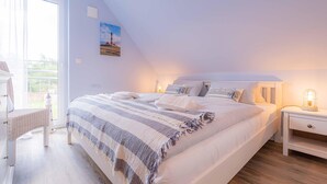 A bedroom with a large bed, a nightstand, a chair, and a window with a view of greenery. - Ferienhaushälfte "zweite Heimat" in Tating für 6 Personen (Tating)