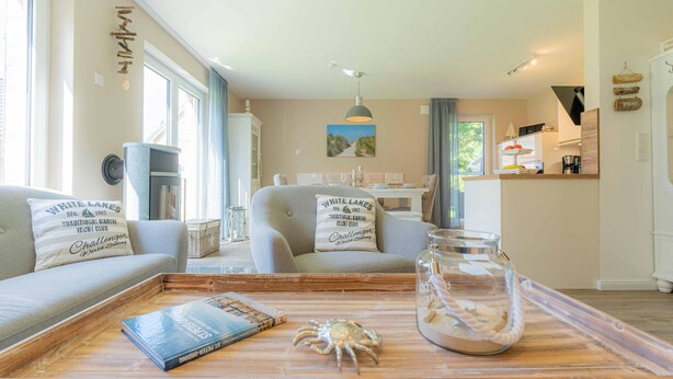 A cozy living room with a sofa, a coffee table with a book and a decorative crab, and a view of the dining area. - Ferienhaushälfte "zweite Heimat" in Tating für 6 Personen (Tating)