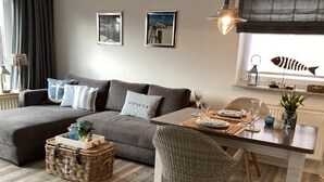 A modern living room with a grey sofa, wicker side table, and a dining area with a white table and wicker chairs. - Appartement Eidernest in Garding für 2 Personen (Garding)