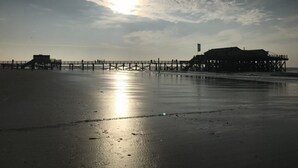 A beach scene with a pier extending into the water, a building on the pier, and the sun setting in the background. - Appartement Eidernest in Garding für 2 Personen (Garding)
