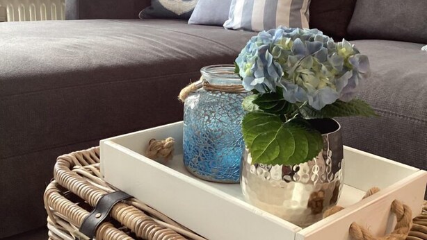 A cozy living room with a grey sofa, blue and white striped pillows, a wicker basket with a tray holding a vase and a lantern. - Appartement Eidernest in Garding für 2 Personen (Garding)