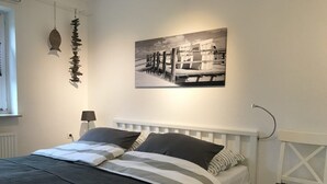 A bedroom with a white bed, a black and white artwork on the wall, a lamp, and a bench. - Appartement Eidernest in Garding für 2 Personen (Garding)