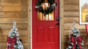 Eine rote Tür mit einem Kranz, zwei kleine Weihnachtsbäume mit Schleifen und einer Fußmatte.