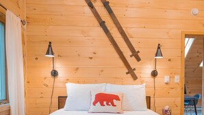 Ein Schlafzimmer mit Holzwandverkleidung, einem Bett, zwei Nachttischlampen, einer Karo-Bettdecke und einem roten Bären-Kissen.