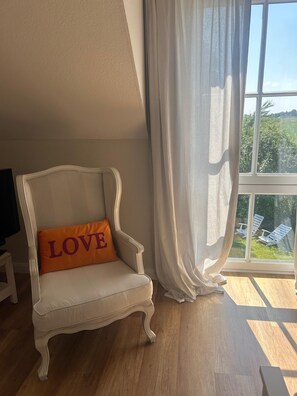 A white armchair with a cushion that has 'LOVE' written on it, positioned next to a window with sheer curtains.