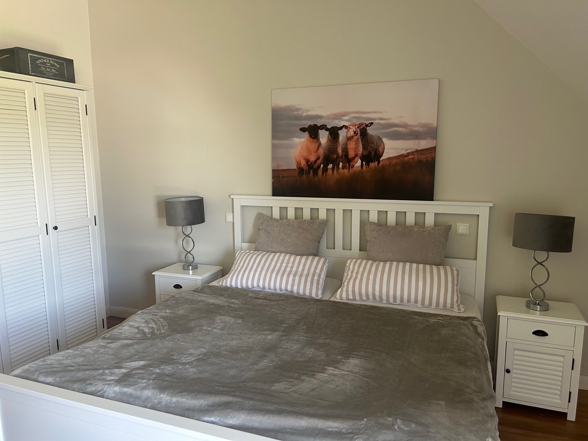 Een slaapkamer met een bed, twee nachtkastjes, een kledingkast en een schilderij van schapen aan de muur.