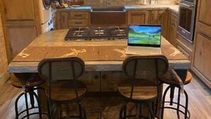 A kitchen with wooden cabinets, a central island with a laptop, hanging light fixtures, and a window with a view. - New! Secluded Cabin w/ Hot Tub, close to Solitude & Brighton Ski Resorts (Brighton)