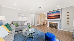 A modern living room with a grey sofa, a glass coffee table, and a blue armchair. There is a fireplace with a painting above it and a kitchen area in the background. - South SJ 4BR: Spacious & Comfy (San Jose)