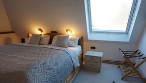 A neatly made bed with a patterned blanket, a bedside table with drawers, a small wooden chair, and a window with a view of the outside.