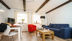 A living room with a blue sofa, a red chair, a wooden coffee table, a white dining table, and a TV. - Ferienwohnung "haubarg Windschuur 4" in Welt für 2 Personen (Welt)