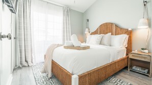 Un dormitorio con una cama de mimbre, ropa de cama blanca, una mesita de noche con lámpara y una alfombra estampada.