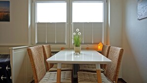 A dining area with a white table, wicker chairs, a potted plant, and a view through window blinds. - Appartement "rochelsand EG" in Tating für 1-3 Personen (Tating)