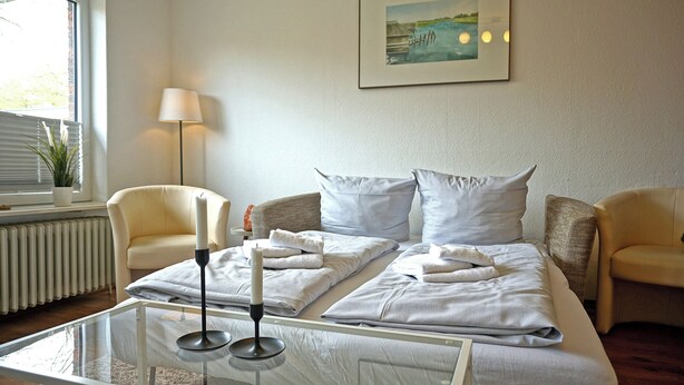 A hotel room with a bed, two chairs, a table with candles, a lamp, a radiator, and a window with blinds. - Appartement "rochelsand EG" in Tating für 1-3 Personen (Tating)