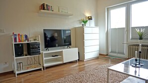 A living room with a TV, bookshelf, white cabinets, and a glass coffee table. - Appartement "rochelsand EG" in Tating für 1-3 Personen (Tating)