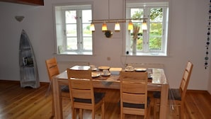 A dining area with a wooden table and chairs, a chandelier, and windows with views of greenery. - Rosenkate in Garding: 130 m² space for 5 people (Garding)