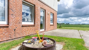A brick building with large windows, a concrete pathway, and a sandbox with toys. - Ferienwohnung auf dem Ferienhof Nickelswarft 2 in Poppenbüll für 2-6 Personen (Poppenbüll)