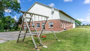 Um edifício com fachada de tijolos e parte superior branca, um conjunto de baloiços e uma área ajardinada com árvores.