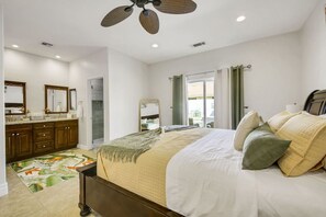 Un dormitorio con cama, baño con tocador, ventilador de techo y vista por la ventana.