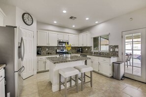 A modern kitchen with white cabinetry, granite countertops, a stainless steel refrigerator, and a central island with bar stools. - Dulce Vida: Pool, Spa, Table Games, Fire Pit, BBQ! (Indio)