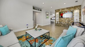Un salon moderne avec une table basse en marbre, un canapé agrémenté de coussins turquoise et un mur décoratif en pierre sur lequel est fixée une télévision diffusant le championnat de la PGA.