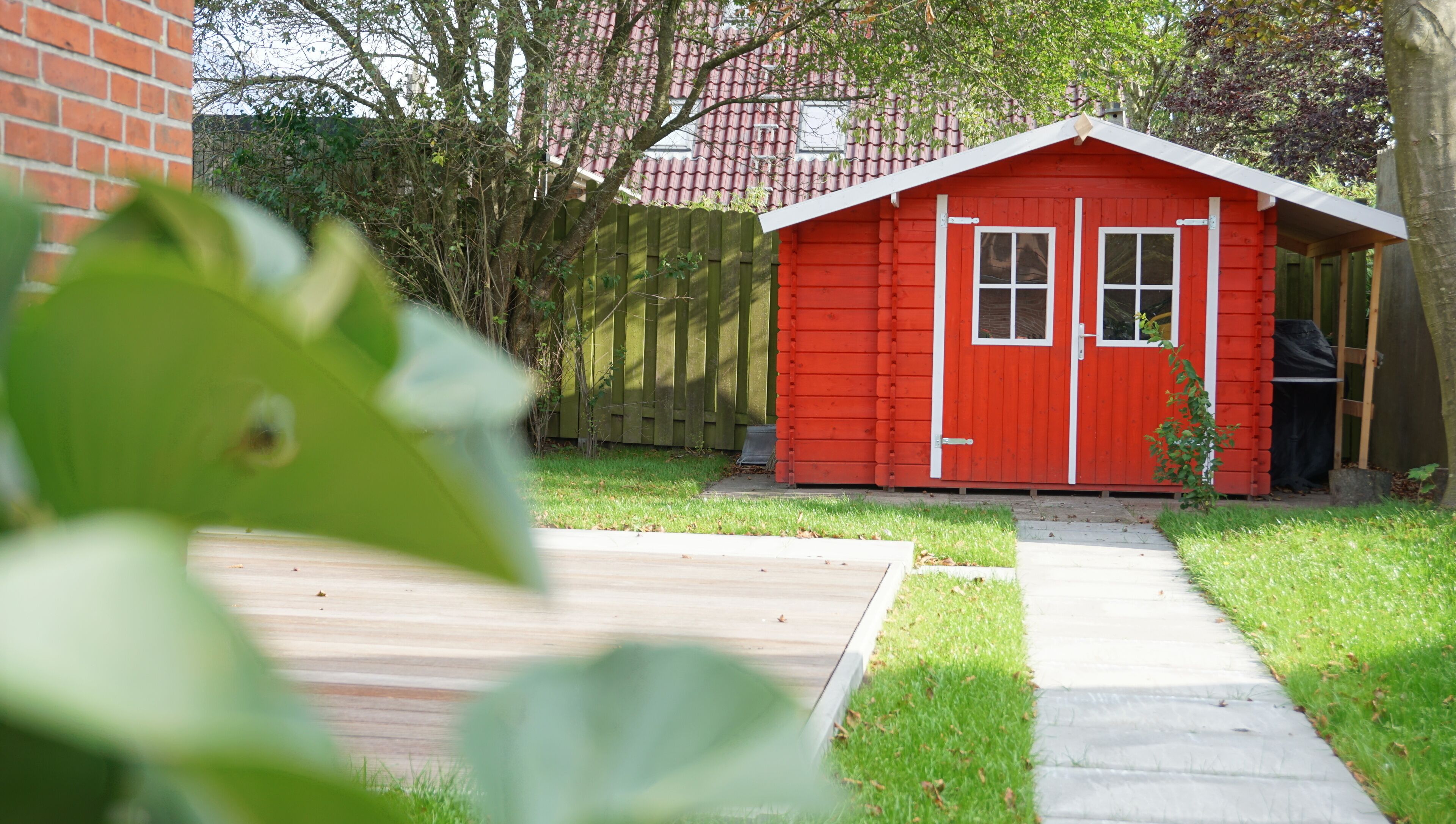 A red wooden shed with a door and windows, surrounded by a grassy yard and a wooden fence.