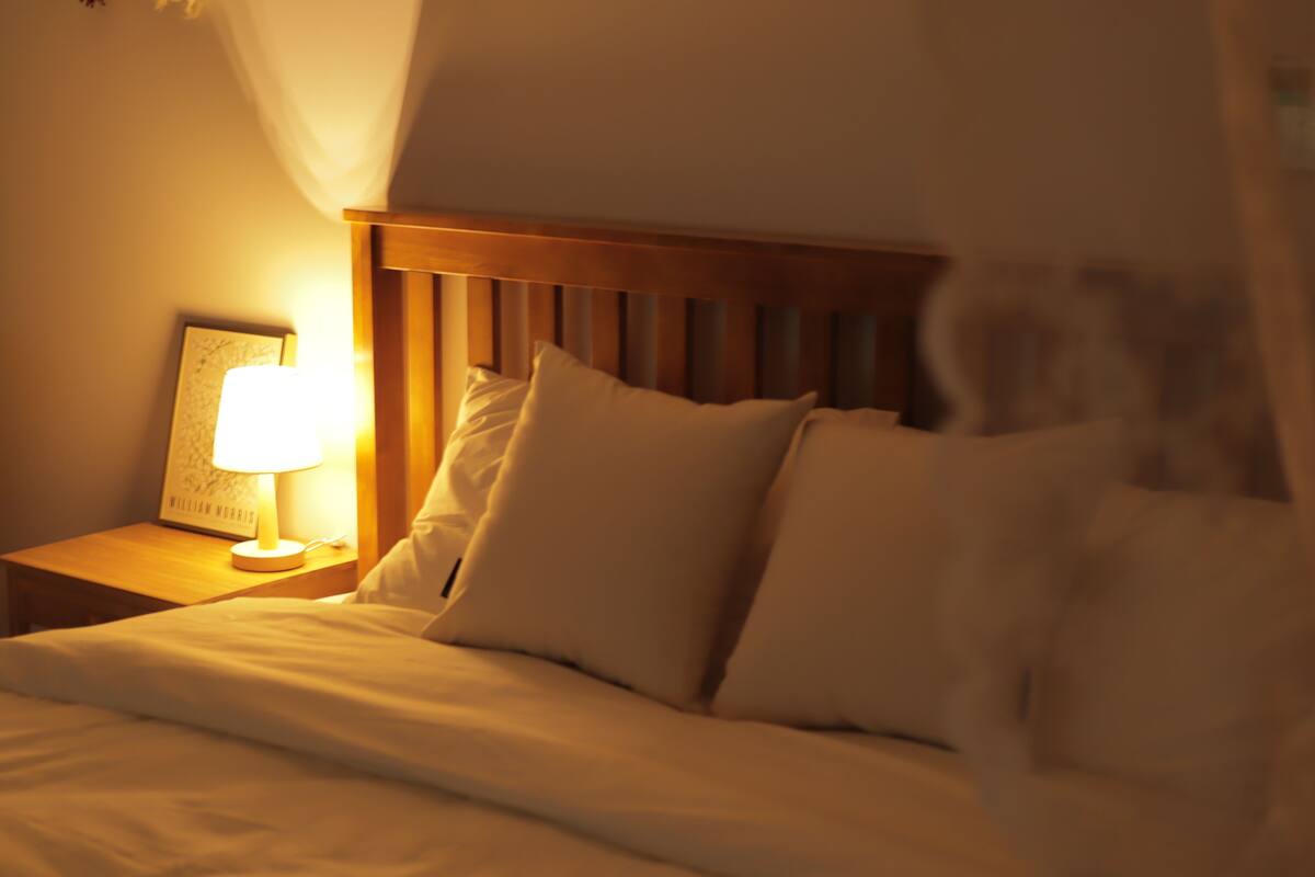 A neatly made bed with white linens and pillows, a bedside table with a lamp, and a framed picture on the wall.