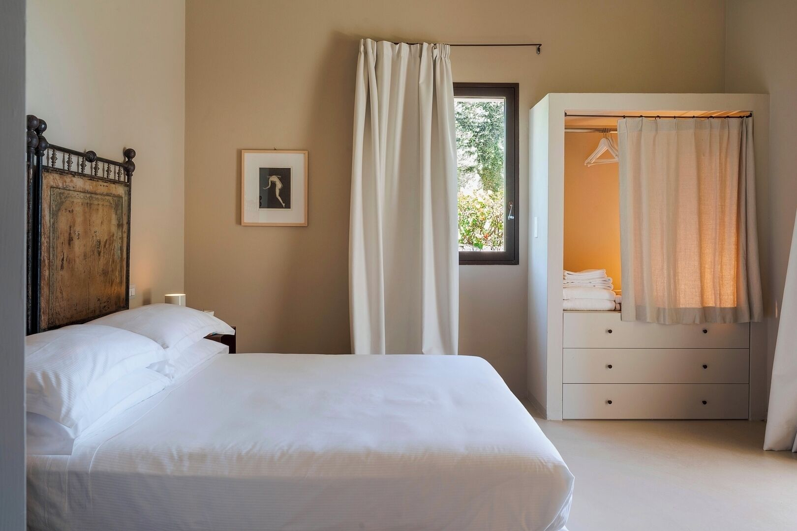 Une chambre à coucher comprenant un lit, une table de chevet, une commode, une fenêtre avec des rideaux et un tableau encadré accroché au mur.