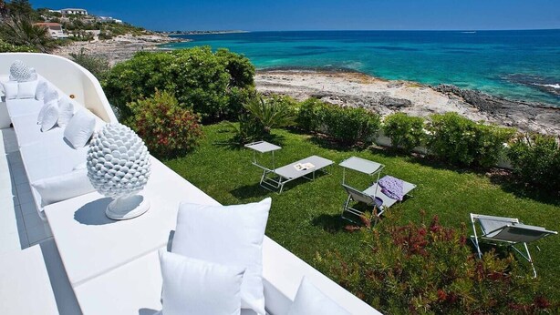 En terrasse med hvide møbler, en dekorativ hvid skulptur og udsigt til havet og den klippefyldte kystlinje.