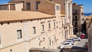 A narrow street with buildings, a clear blue sky, and a flower pot with orange flowers in the foreground. - Casa Dei Nonni - Apartment in Syracuse (Syracuse)