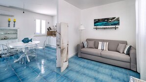 Een moderne woonkamer met een grijze bank, een eettafel met transparante stoelen en een keuken met witte kasten.