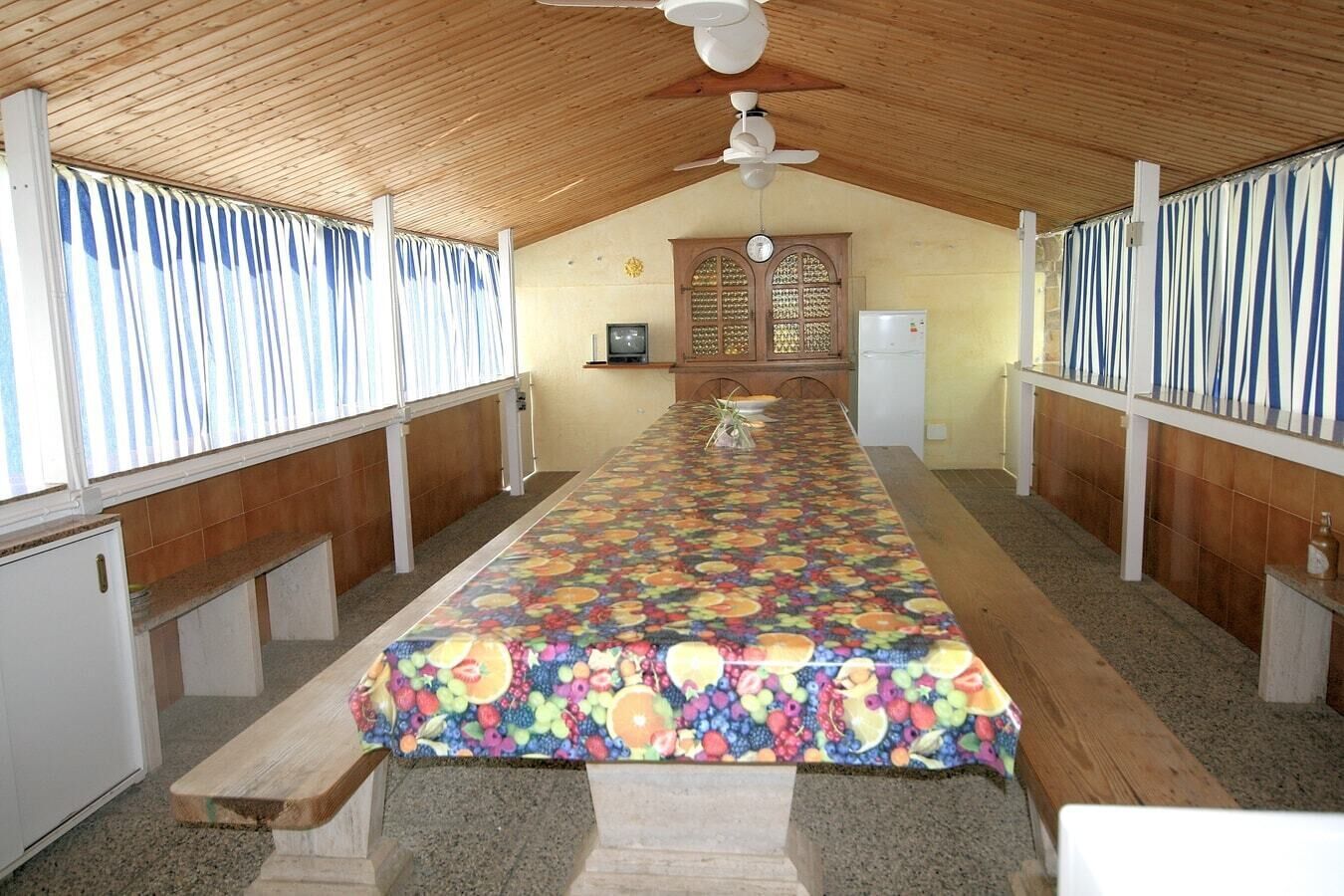 A long, narrow room with a wooden ceiling and a ceiling fan. There is a long table with a colorful tablecloth, and a small refrigerator and sink are visible in the background.