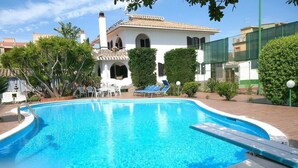 A swimming pool with lounge chairs and a patio area, surrounded by greenery and a building with a tiled roof. - Villa Trapezio with private pool and tennis court (Quartu Santa Elena)