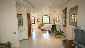 A spacious living room with a large window, a sofa, a dining table, and a television. - Villa Trapezio with private pool and tennis court (Quartu Santa Elena)