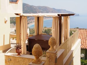 A balcony with a wooden table and chairs, a potted plant, and a view of the sea and mountains.