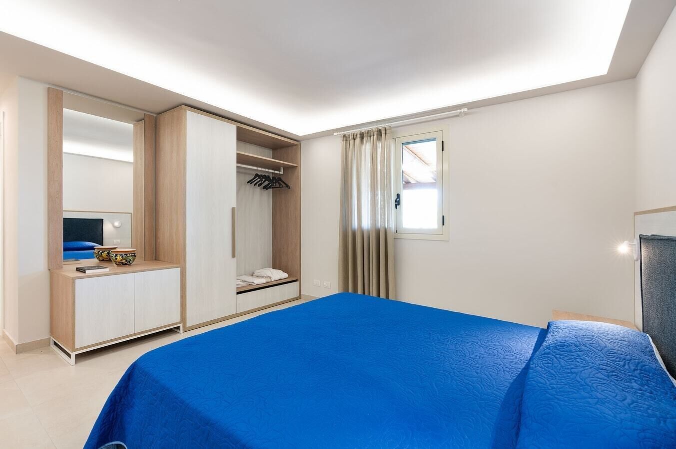 Une chambre d’hôtel avec un lit, une armoire, un miroir et une fenêtre avec des rideaux.
