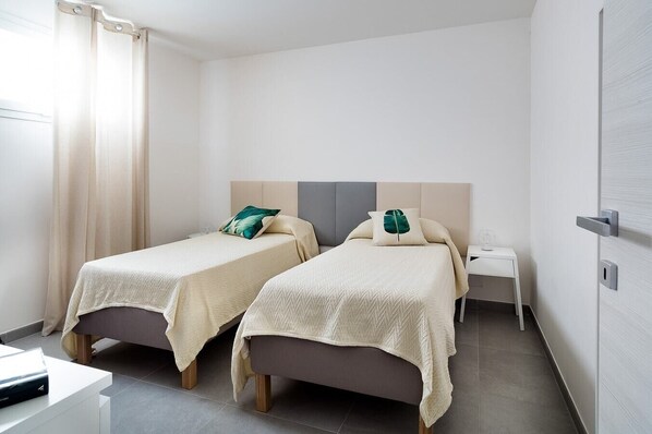 A modern hotel room with two single beds, a bedside table with a glass, and a door with a handle.
