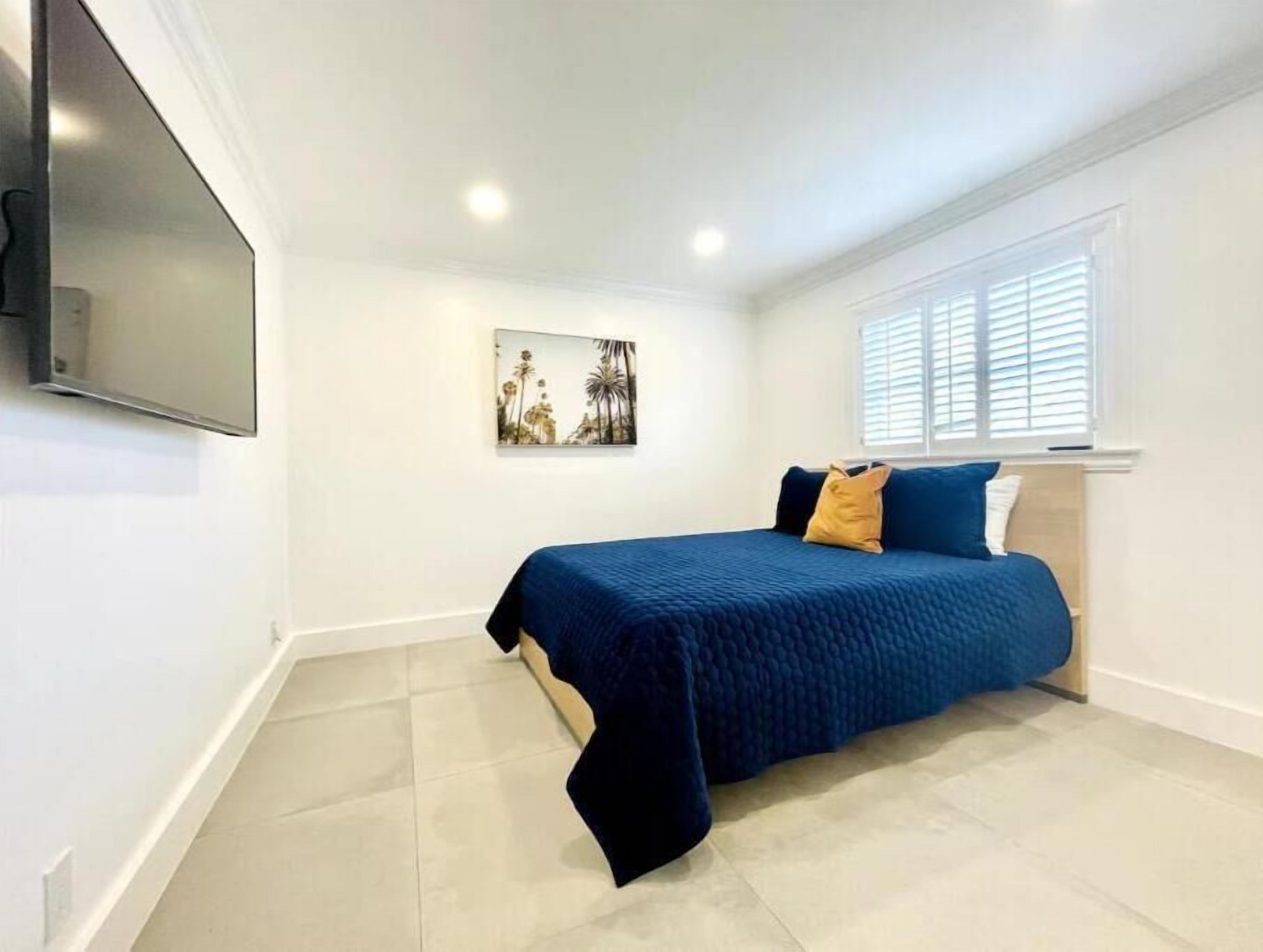 A bedroom with a bed, a wall-mounted TV, a window with blinds, and a painting on the wall.