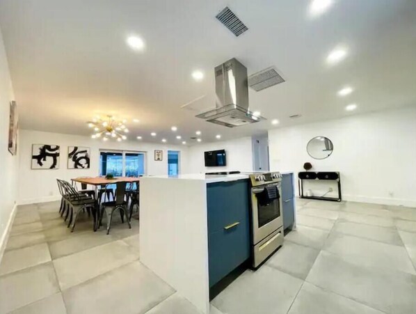 A modern kitchen with a central island, built-in oven, and microwave. There is a dining area with chairs and a table, and a large window providing natural light.