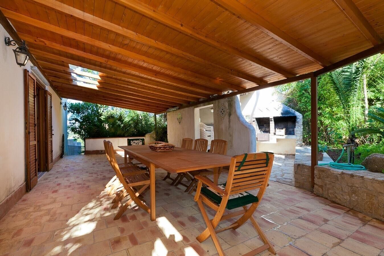 An outdoor patio with a wooden table and chairs, surrounded by stone tiles and a wooden ceiling.