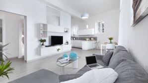 Ein modernes Wohnzimmer mit einem grauen Sofa, einem Glastisch und einem Fernseher. Es gibt eine Küchenzeile mit weißen Schränken und einem Esstisch.