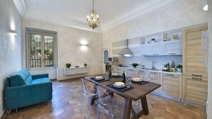 Una cuina moderna amb armaris blancs, una zona de menjador amb taula i cadires de fusta, i un sofà blau.
