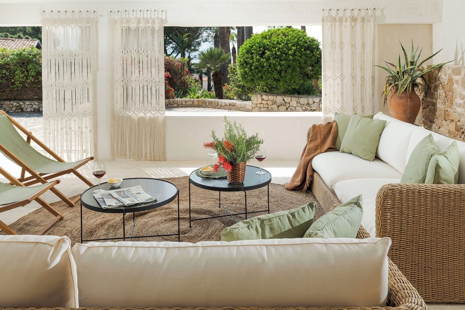 A cozy outdoor seating area with a wicker sofa, a round table, and a chair, surrounded by white curtains and a view of greenery.