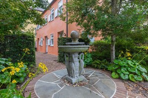 Un jardín con una fuente de piedra, abundante follaje y un edificio al fondo.