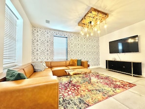 Un salon moderne avec un canapĂ© dâangle, un tapis Ă motifs, un luminaire au plafond et un tĂ©lĂ©viseur Ă Ă©cran plat fixĂ© au mur.