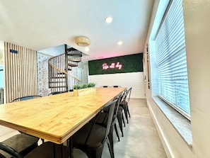 Un espace repas avec une table et des chaises en bois, un escalier, un panneau lumineux au nĂ©on et une fenĂȘtre avec des stores.