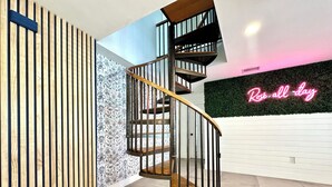 Una escalera de caracol con barandilla de madera y pasamanos de metal negro, una pared con listones verticales de madera, y un letrero de neón que dice 'Rose-all-day'.