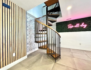 Un escalier en colimaçon avec une main courante en bois et des rampes en métal noir, un mur avec des lattes de bois verticales, et un panneau néon sur lequel on peut lire « Rose-all-day ».