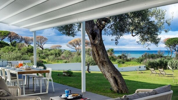 A covered outdoor dining area with a table set for two, overlooking a grassy landscape and the sea.