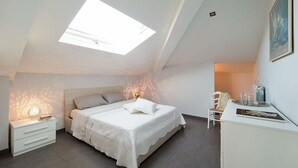 A bedroom with a skylight, a bed with white bedding, a nightstand, a chair, and a desk with a lamp.